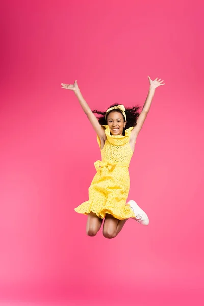 Full length view of happy curly african american child in yellow outfit jumping with hands in air on pink background — Stock Photo