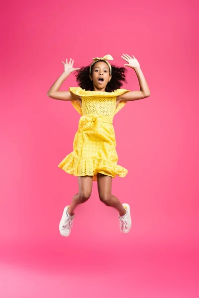 Vista completa de niño afroamericano rizado impactado en traje amarillo saltando sobre fondo rosa - foto de stock