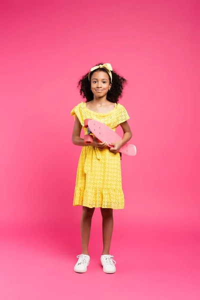 Vue pleine longueur de sourire frisé afro-américain enfant en tenue jaune avec penny board sur fond rose — Photo de stock