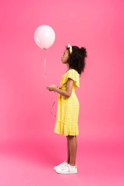 Vista lateral del niño afroamericano rizado en traje amarillo con globo sobre fondo rosa - foto de stock