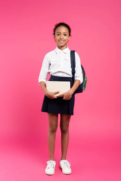 Vista frontal da colegial afro-americana sorridente com mochila e livros sobre fundo rosa — Fotografia de Stock