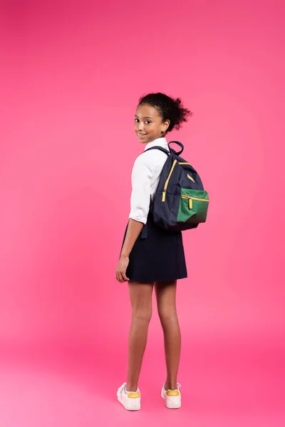 Rückansicht eines lächelnden afrikanisch-amerikanischen Schulmädchens mit Rucksack auf rosa Hintergrund — Stockfoto