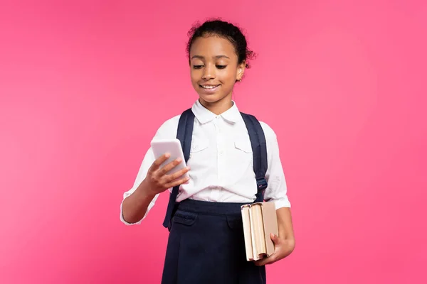 Colegiala afroamericana sonriente con libros y teléfono inteligente aislado en rosa - foto de stock