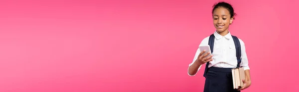 Smiling african american schoolgirl with books and smartphone isolated on pink, panoramic shot — Stock Photo