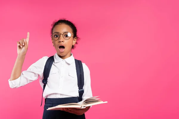 Sorprendido afroamericano colegiala en gafas celebración libro y mostrando idea gesto aislado en rosa - foto de stock