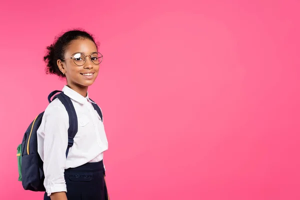 Colegiala afroamericana sonriente en gafas con mochila aislada en rosa - foto de stock
