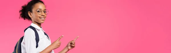 Smiling african american schoolgirl in glasses with backpack pointing aside isolated on pink, panoramic shot — Stock Photo