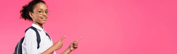 Sorridente studentessa afroamericana in occhiali con zaino a parte isolato sul rosa, scatto panoramico — Foto stock