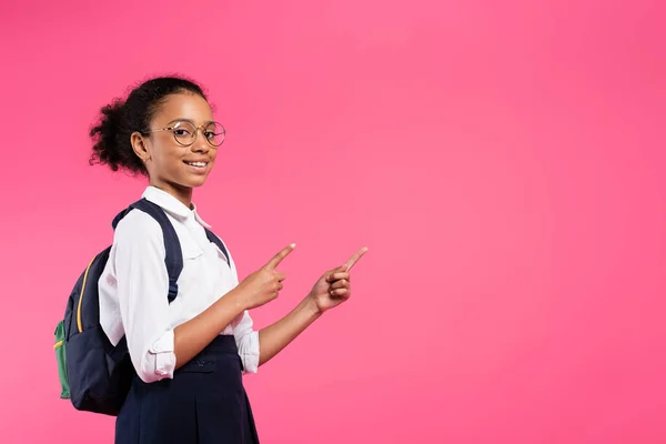 Sonriente afroamericana colegiala en gafas con mochila apuntando a un lado aislado en rosa - foto de stock