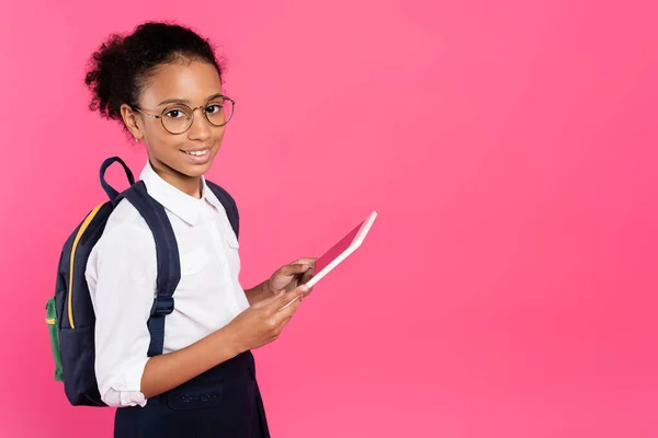 Smiling african american schoolgirl in glasses with backpack using digital tablet isolated on pink — Stock Photo