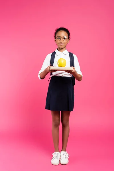 Smiling african american schoolgirl in glasses with book and apple on pink — Stock Photo