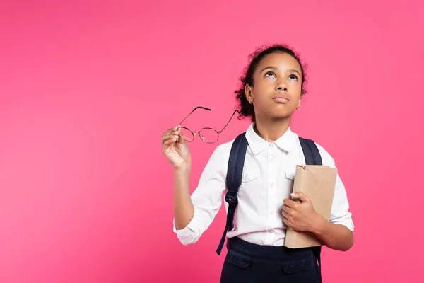 Пенсійна афроамериканська школярка з книгою та окулярами, яка дивиться вгору ізольовано на рожевому — стокове фото