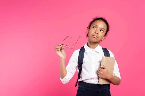 Pensive african american schoolgirl with book and glasses isolated on pink — Stock Photo