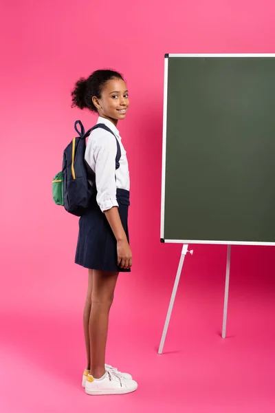 Sonriente afroamericana colegiala con mochila cerca de pizarra vacía en rosa — Stock Photo