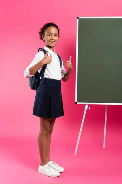 Smiling african american schoolgirl with backpack near empty chalkboard showing thumbs up on pink background — Stock Photo