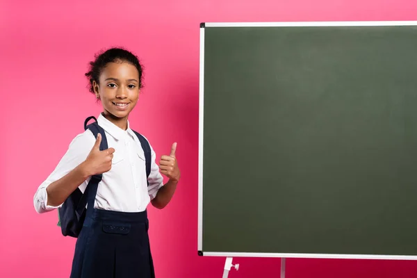 Sonriente afroamericana colegiala con mochila cerca de pizarra vacía mostrando pulgares hacia arriba sobre fondo rosa - foto de stock