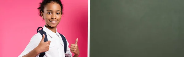 Smiling african american schoolgirl with backpack near empty chalkboard showing thumbs up on pink background, panoramic shot — Stock Photo