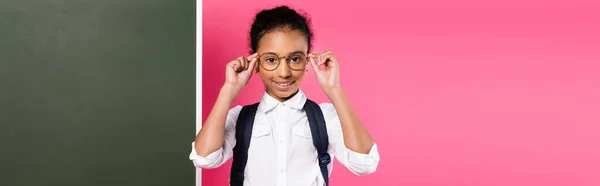 Smiling african american schoolgirl with backpack near empty chalkboard on pink background, panoramic shot — Stock Photo