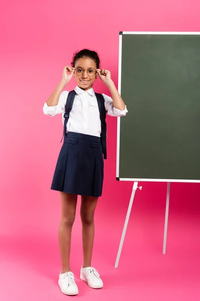 Smiling african american schoolgirl with backpack near empty chalkboard on pink background — Stock Photo