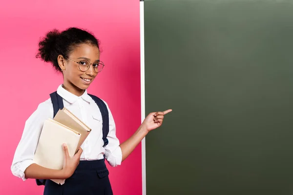 Smiling african american schoolgirl with books pointing at empty chalkboard on pink background — Stock Photo