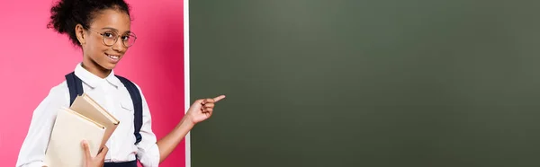 Smiling african american schoolgirl with books pointing at empty chalkboard on pink background, panoramic shot — Stock Photo