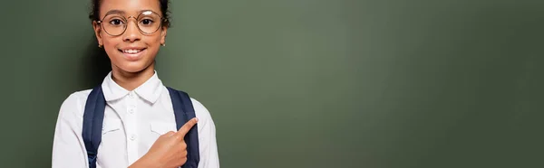 Smiling african american schoolgirl pointing at empty green chalkboard, panoramic shot — Stock Photo