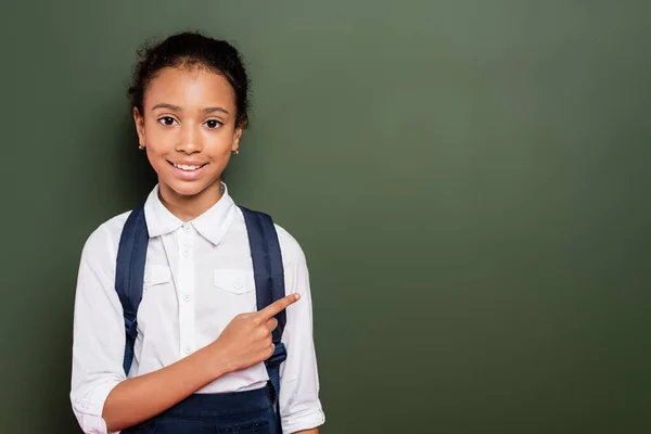 Smiling african american schoolgirl pointing at empty green chalkboard — Stock Photo