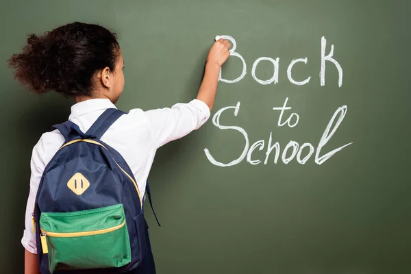 Vista posterior de la colegiala afroamericana con mochila escribiendo de nuevo a la inscripción de la escuela en pizarra verde - foto de stock