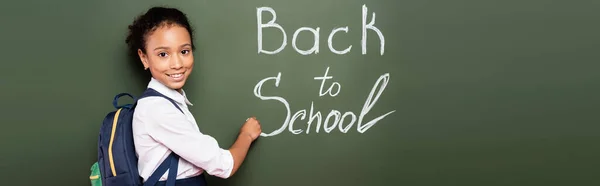 Sonriente colegiala afroamericana con mochila escribiendo de nuevo a la inscripción de la escuela en pizarra verde, tiro panorámico - foto de stock