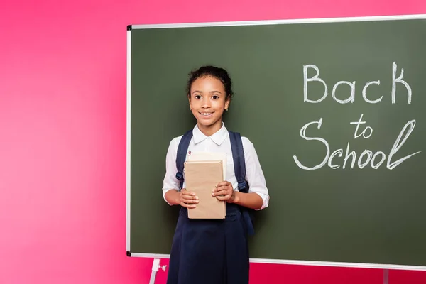 Smiling african american schoolgirl with books near back to school inscription on green chalkboard on pink background — Stock Photo