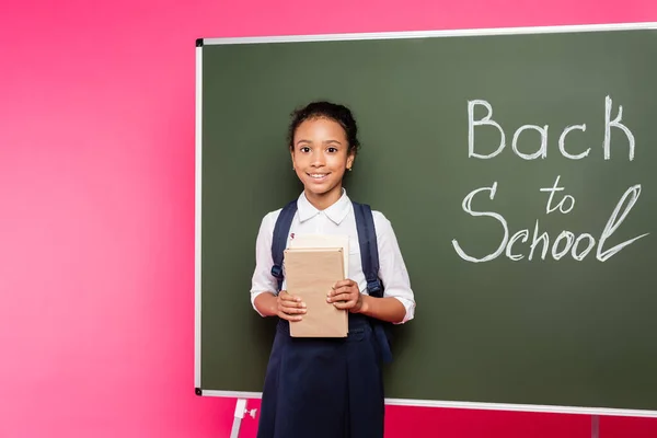 Souriant afro-américaine écolière avec des livres près de retour à l'école inscription sur tableau vert sur fond rose — Photo de stock