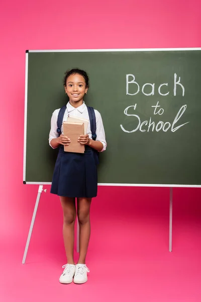 Smiling african american schoolgirl with books near back to school inscription on green chalkboard on pink background — Stock Photo
