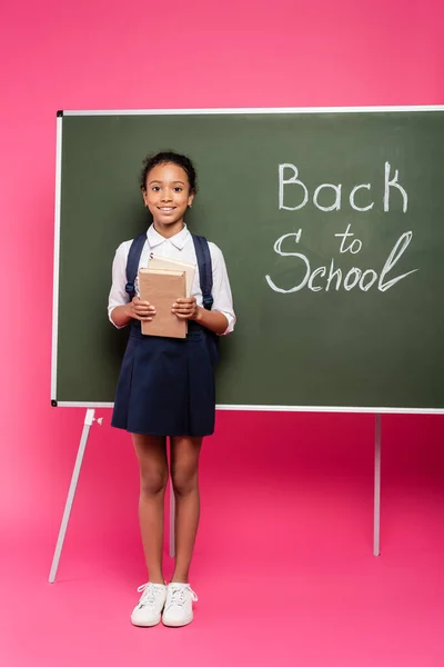 Lächelndes afrikanisch-amerikanisches Schulmädchen mit Büchern in der Nähe der Schulinschrift auf grüner Kreidetafel auf rosa Hintergrund — Stockfoto