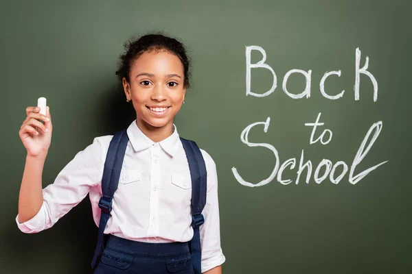 Smiling african american schoolgirl with chalk near back to school inscription on green chalkboard — Stock Photo