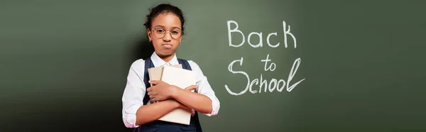 Upset african american schoolgirl with books near back to school inscription on green chalkboard, panoramic shot — Stock Photo