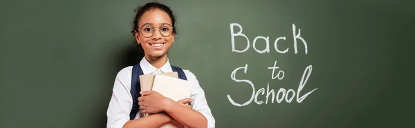 Усміхнена афроамериканська школярка з книгами біля школи на зеленій дошці, панорамний знімок — стокове фото