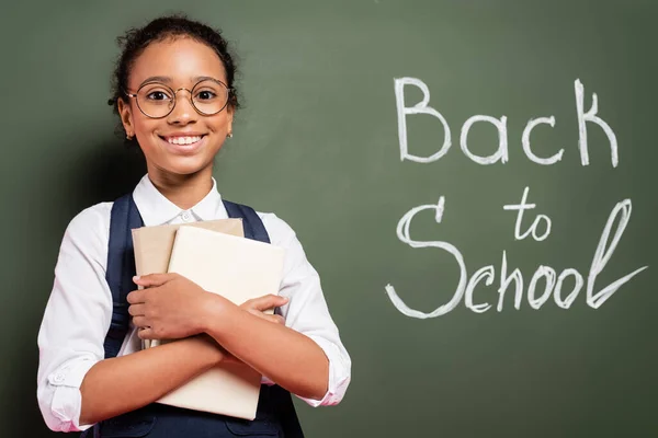 Scolaretta afro-americana sorridente con libri vicino all'iscrizione scolastica sulla lavagna verde — Foto stock