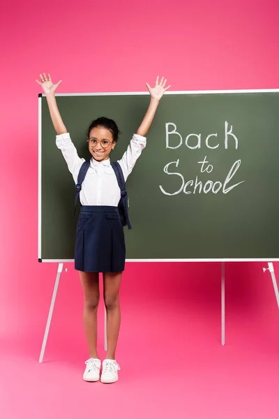 Aluna americana africana feliz em óculos com as mãos no ar perto de volta à inscrição da escola no quadro verde no fundo rosa — Fotografia de Stock