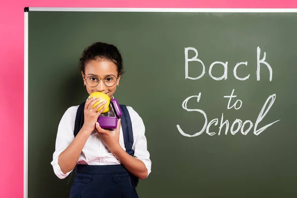 Sonriente afroamericana colegiala en gafas comiendo manzana cerca de vuelta a la escuela inscripción en pizarra verde sobre fondo rosa - foto de stock