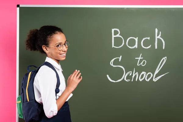 Smiling african american schoolgirl in glasses near back to school inscription on green chalkboard on pink background — Stock Photo