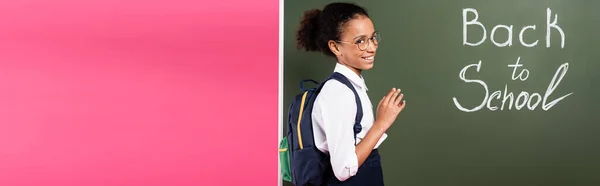 Souriant afro-américaine écolière dans des lunettes près de retour à l'école inscription sur tableau vert sur fond rose, vue panoramique — Photo de stock