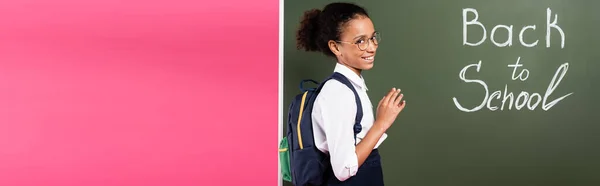 Smiling african american schoolgirl in glasses near back to school inscription on green chalkboard on pink background, panoramic shot — Stock Photo