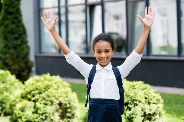 Smiling african american schoolgirl with backpack and hands in air outdoors — Stock Photo