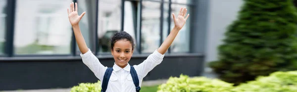 Smiling african american schoolgirl with backpack and hands in air outdoors, panoramic shot — Stock Photo