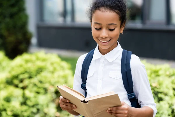 Smiling african american schoolgirl reading book outdoors — Stock Photo