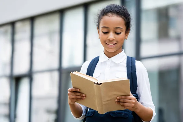 Souriant afro-américaine écolière lecture livre en plein air — Photo de stock