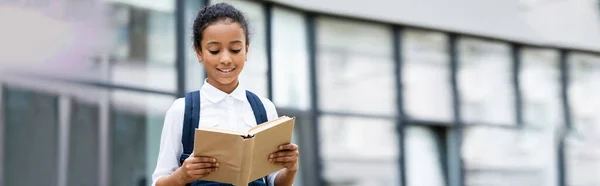 Smiling african american schoolgirl reading book outdoors, panoramic shot — Stock Photo