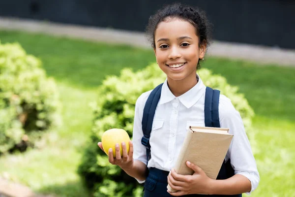 Smiling african american schoolgirl with book and apple outdoors — Stock Photo