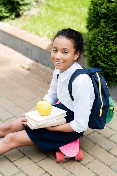 Sonriente afroamericano colegiala con libros y manzana sentado en penny board - foto de stock