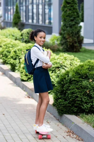Glücklich afrikanisch-amerikanische Schulmädchen mit Rucksack und Bücher Reiten Penny Board im Freien — Stockfoto